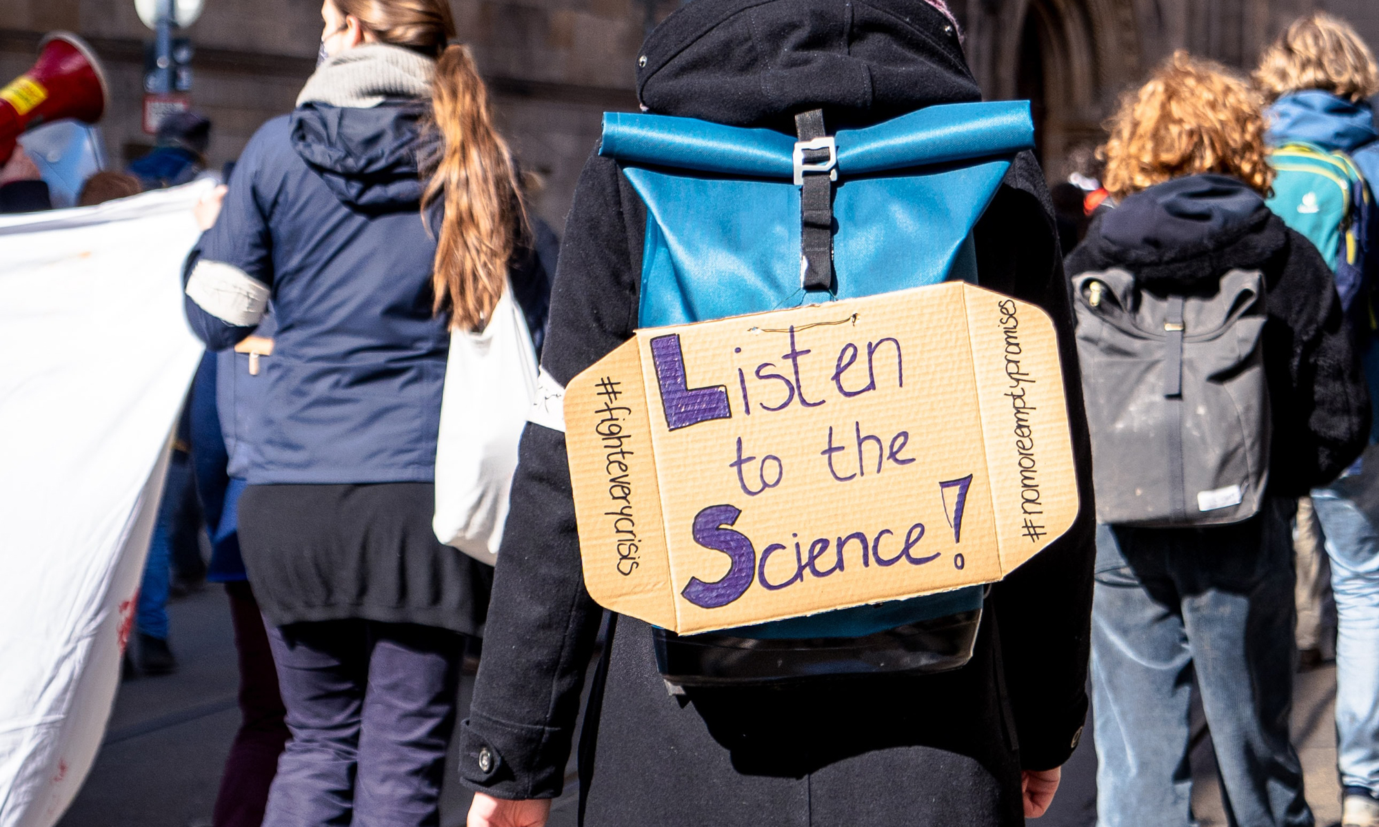 Protest showing people demonstrating. One sign in the centre shows the words "Listen to Science". Photo by Mika Baumeister on Unsplash